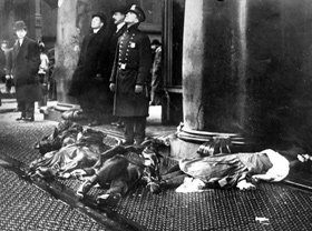 A police officer and others look up in shock at workers poised to jump from the upper floors of the burning Asch Building with the broken bodies of Triangle fire victims at their feet